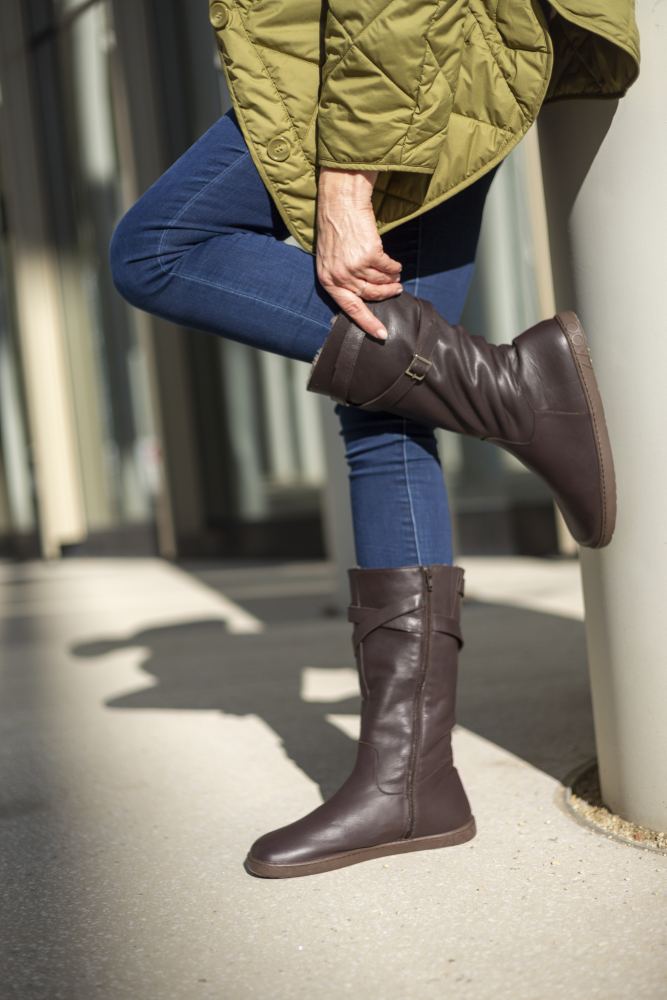 Eine Person in Blue Jeans und ATTRAQT Winter Brown-Stiefeln von ZAQQ lehnt an einem Pfosten und hat ein Bein über das andere geschlagen. Sie trägt eine grüne Steppjacke und genießt den Komfort ihres Outfits, während im Hintergrund Sonnenlicht durch die Glasfenster fällt.