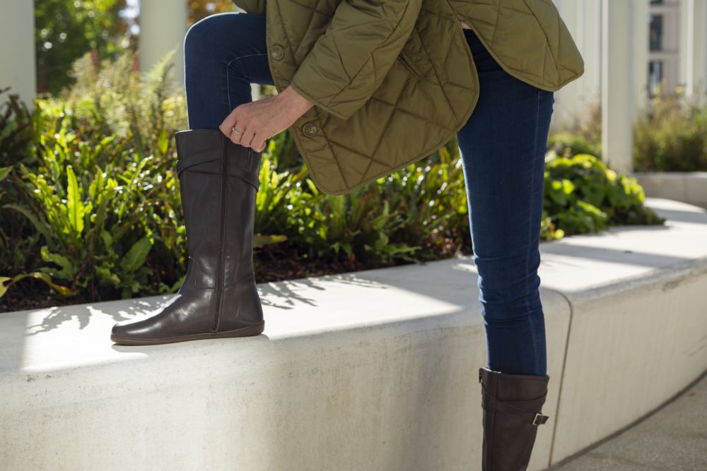 Eine Person in einer grünen Steppjacke richtet ihre kniehohen ATTRAQT Nappa Brown-Stiefel von ZAQQ, während sie an einer niedrigen Betonmauer lehnt. Sie sind an einem sonnigen Tag im Freien mit grünem Laub im Hintergrund.