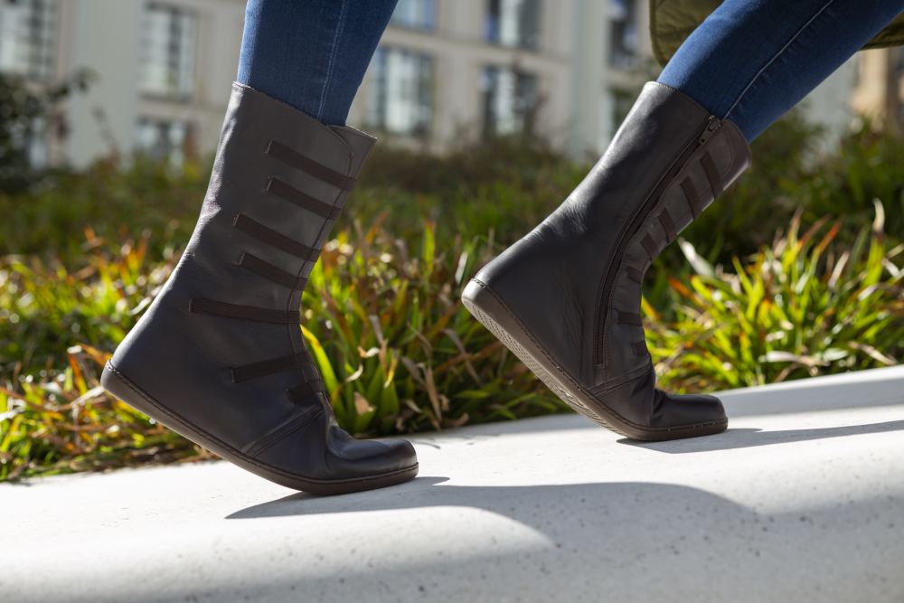 Nahaufnahme einer Person, die in ZAQQs ATTRAQT Stretch Nappa Brown-Stiefeln und Blue Jeans auf einem weißen Felsvorsprung läuft. Das satte braune Leder der Stiefel strahlt Stil und Flexibilität aus, während grünes Laub und verschwommene Gebäude im Hintergrund eine städtische Umgebung suggerieren.