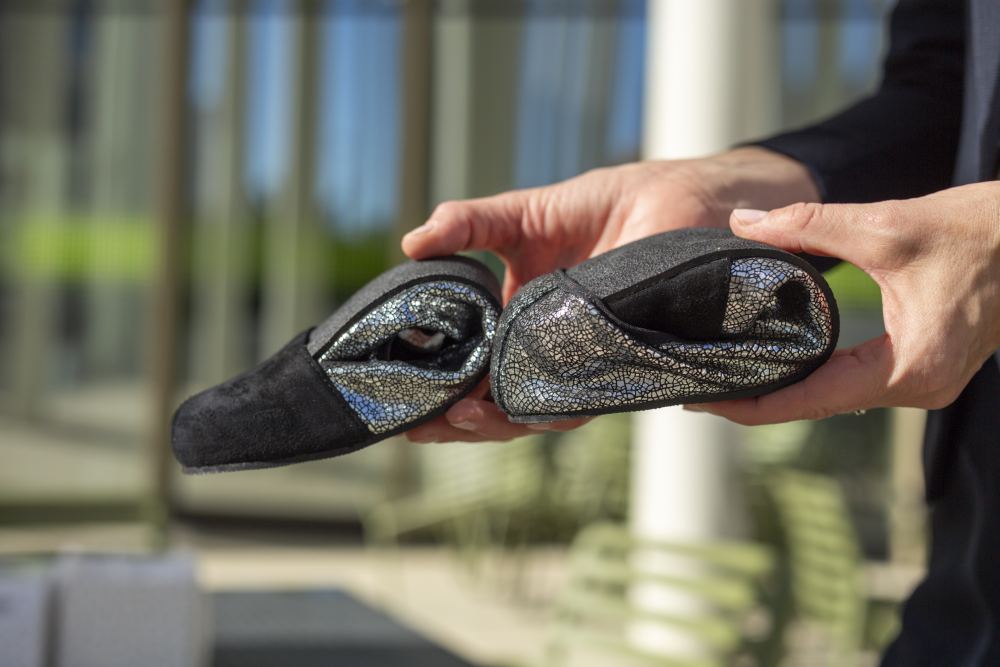 Eine Person hält den TWIST Derby Black, ein Paar flexible Slipper von ZAQQ, mit glänzenden, strukturierten Details, die an Veloursleder erinnern. Die Schuhe sind gefaltet, um ihre Weichheit und Tragbarkeit vor einem verschwommenen Outdoor-Hintergrund mit einem Hauch von Grün zu präsentieren.