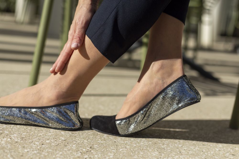 Eine Person sitzt im Freien und berührt ihren Knöchel. Dabei kommen die glitzernden TWIST Derby Black Flats von ZAQQ zur Geltung. Im Hintergrund sind verschwommene grüne Stühle und Tische zu sehen, auf deren Boden das Sonnenlicht verspielte Schatten wirft.