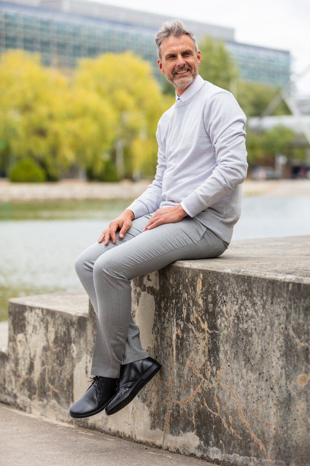 Ein Mann mit grauem Haar und Bart sitzt auf einem Betonvorsprung am Flussufer. Er trägt einen hellgrauen Pullover und eine Hose sowie schwarze DUQE-Stiefel von ZAQQ. Er lächelt und blickt an einem bewölkten Tag in die Ferne, mit Bäumen und einem Gebäude im Hintergrund.