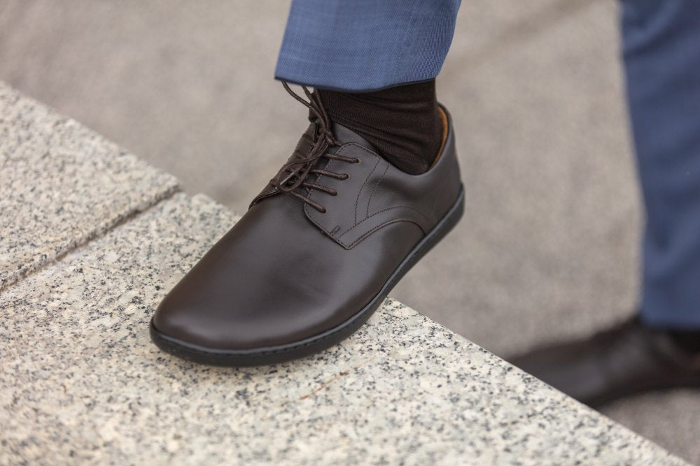 Eine Person, die ein Paar dunkelbraune PEAQ-Schuhe von ZAQQ und eine blaue Hose trägt, betritt im mühelosen Business-Casual-Stil eine Steinoberfläche.