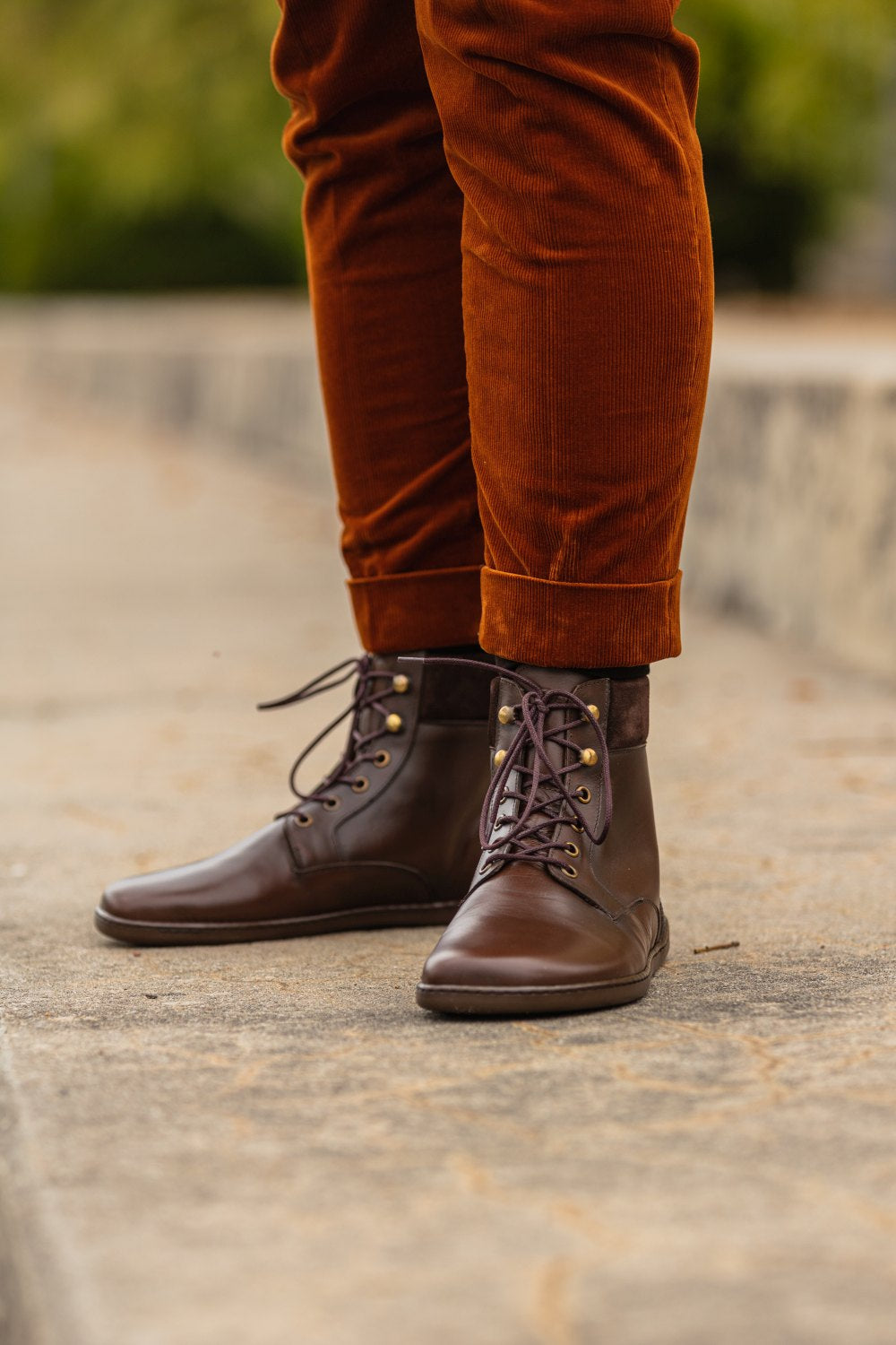 Eine Person trägt stylische QORBY Brown Boots von ZAQQ, also halbhohe Stiefel mit Schnürsenkeln, dazu hochgekrempelte rostfarbene Hosen und steht auf einem Gehsteig. Die Umgebung ist leicht verschwommen, was auf eine Außenkulisse hindeutet.