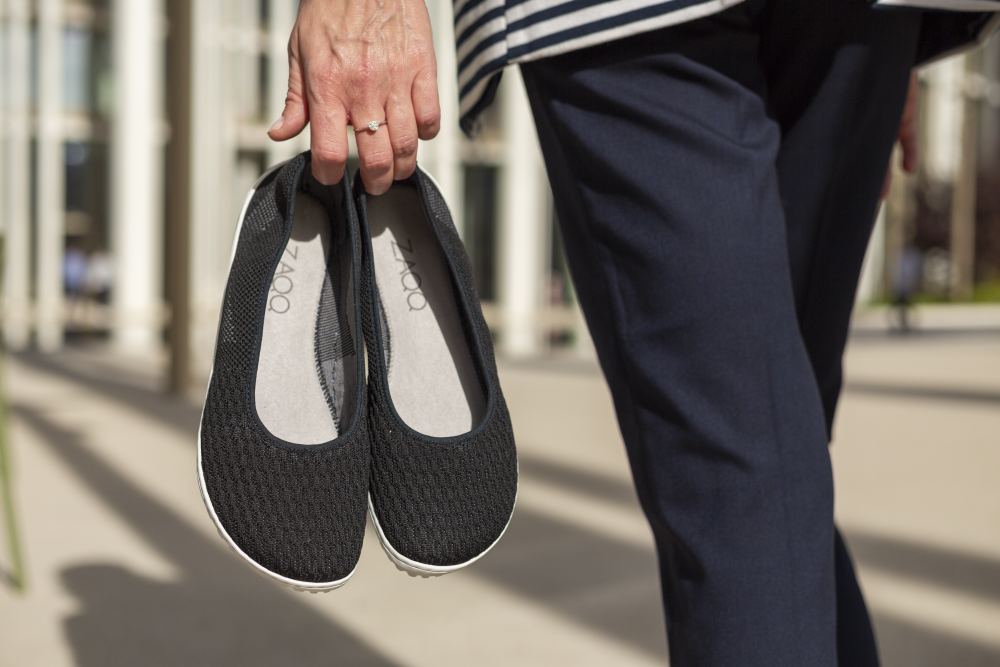 Eine Person in marineblauer Hose und gestreiftem Oberteil steht an einem sonnigen Tag im Freien und hält schwarze Slipper von SUQAR von ZAQQ in der Hand. Jeder Schuh hat eine einzigartige Sneakersohle für den bequemen, femininen Look.