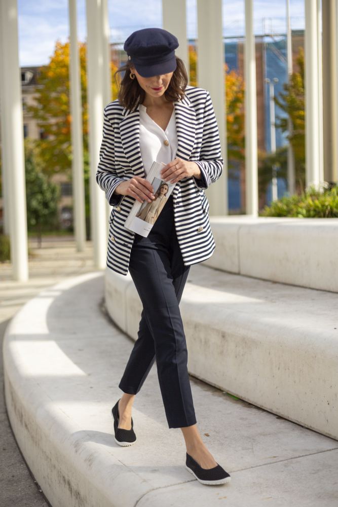 Eine Frau in gestreiftem Blazer, weißer Bluse und schwarzer Hose geht mit einer Zeitschrift in der Hand nach draußen. Sie trägt eine marineblaue Kappe und schwarze SUQAR-Ballerinas von ZAQQ und strahlt inmitten der sonnigen Stadtkulisse mit Bäumen und moderner Architektur im Hintergrund einen eleganten, femininen Look aus.