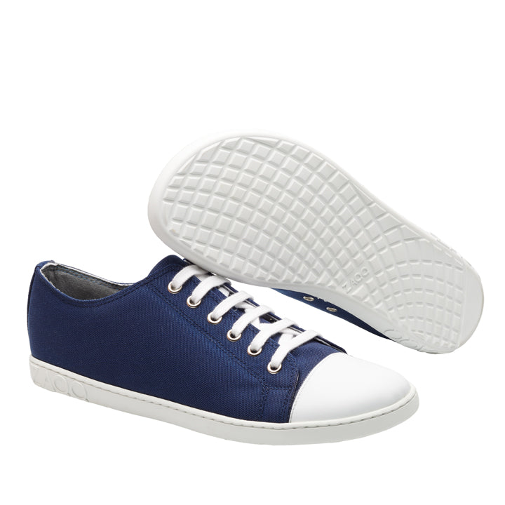 Wir präsentieren die CHUQQS Low Blue Sneaker von ZAQQ, die eine Kombination aus marineblauem Canvas und weißen Zehenkappen aus Leder bieten. Diese bequemen Schuhe sind mit weißen Schnürsenkeln und Sohlen gestaltet. Ein Schuh steht aufrecht, während der andere auf der Seite liegt und eine schicke Sohle mit Gittermuster aufweist, die ideal für Barfußschuh-Fans ist.