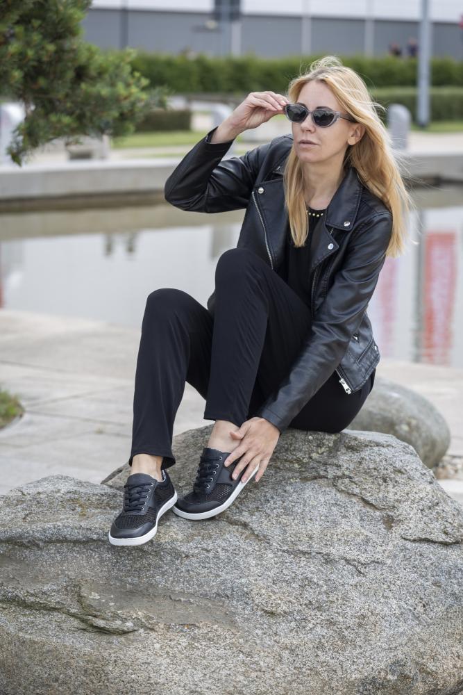 Eine Person mit langen blonden Haaren, einer Sonnenbrille ALIQANTE Black von ZAQQ und einer schwarzen Lederjacke sitzt auf einem großen Felsen im Freien. Sie rückt ihre Sonnenbrille mit einer Hand zurecht und präsentiert dabei ihren Barfußschuh-Stil, unter dem schwarze Turnschuhe hervorschauen.