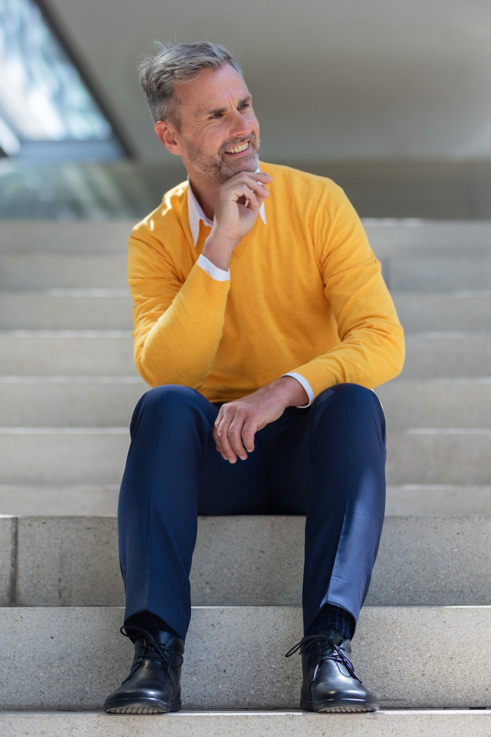 Ein lächelnder Mann mit grauem Haar und Bart sitzt auf einer Treppe und strahlt in seinem leuchtend gelben Pullover, den blauen Hosen und den stylischen schwarzen ZAQQ ARQ-Schuhen Eleganz aus. Seine rechte Hand ruht nachdenklich auf seinem Kinn, während er vor einem leicht verschwommenen Hintergrund zur Seite blickt.