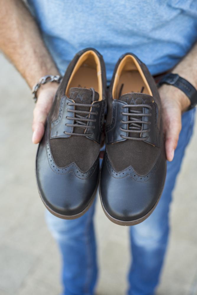 Eine Person hält ein Paar stylische BRIQ Brogue Tonka Schuhe von ZAQQ, gefertigt aus dunkelbraunem Glattleder und Wildleder mit klassischen Brogue-Details. Sie trägt ein blaues T-Shirt und Jeans, die diese eleganten Business-Schuhe perfekt ergänzen.