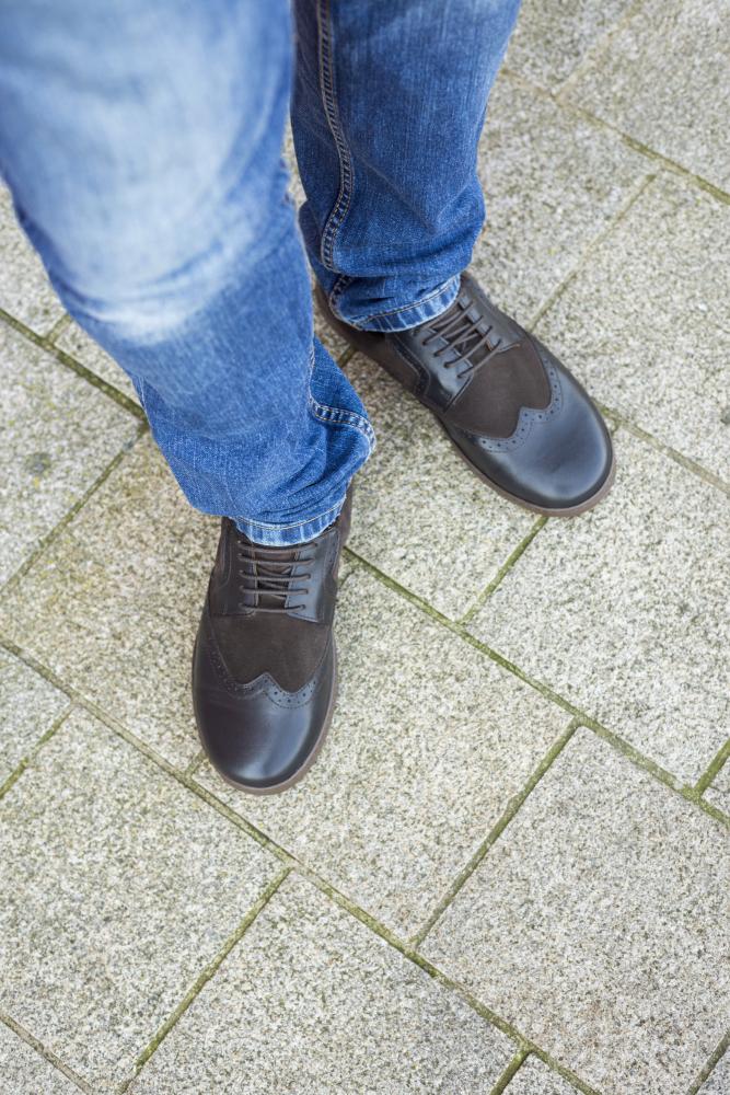 Eine Person in Blue Jeans steht auf einem Steinpflaster und trägt elegante BRIQ Brogue Tonka von ZAQQ, gefertigt aus schwarzem Leder und Wildleder.