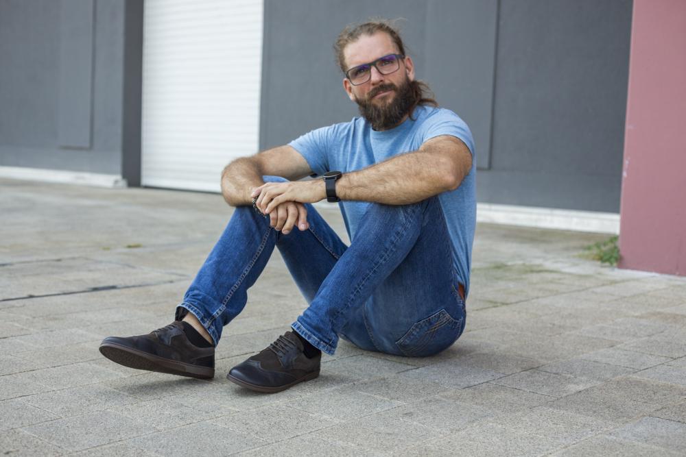 Ein bärtiger Mann mit Brille und Pferdeschwanz sitzt lässig auf dem Bürgersteig. Er trägt ein hellblaues T-Shirt, blaue Jeans und dunkle BRIQ Brogue Tonka-Schuhe von ZAQQ. Seine Arme ruhen auf seinen Knien und er blickt vor einem Hintergrund aus grauen und rosa Wänden in die Kamera.