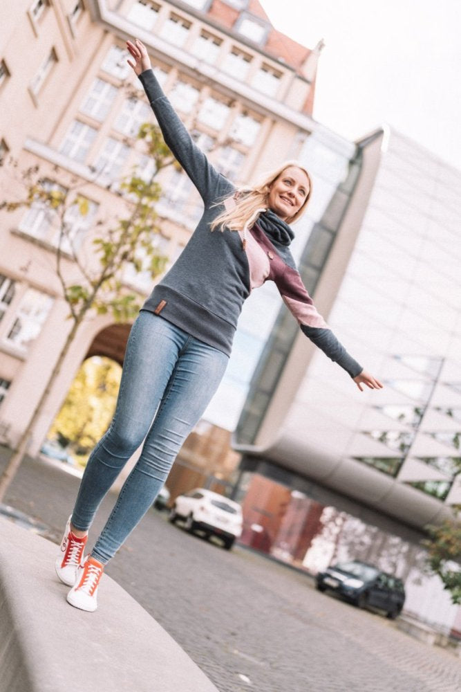 Eine Frau in grauem Pullover und Bluejeans balanciert spielerisch auf einer niedrigen Mauer inmitten einer urbanen Kulisse aus modernen und historischen Gebäuden. Sie trägt leuchtend rote CEDRIQ-Sneaker von ZAQQ mit Kautschuk-Sohlen und lächelt, während sie ihre Arme ausstreckt, um das Gleichgewicht zu halten.