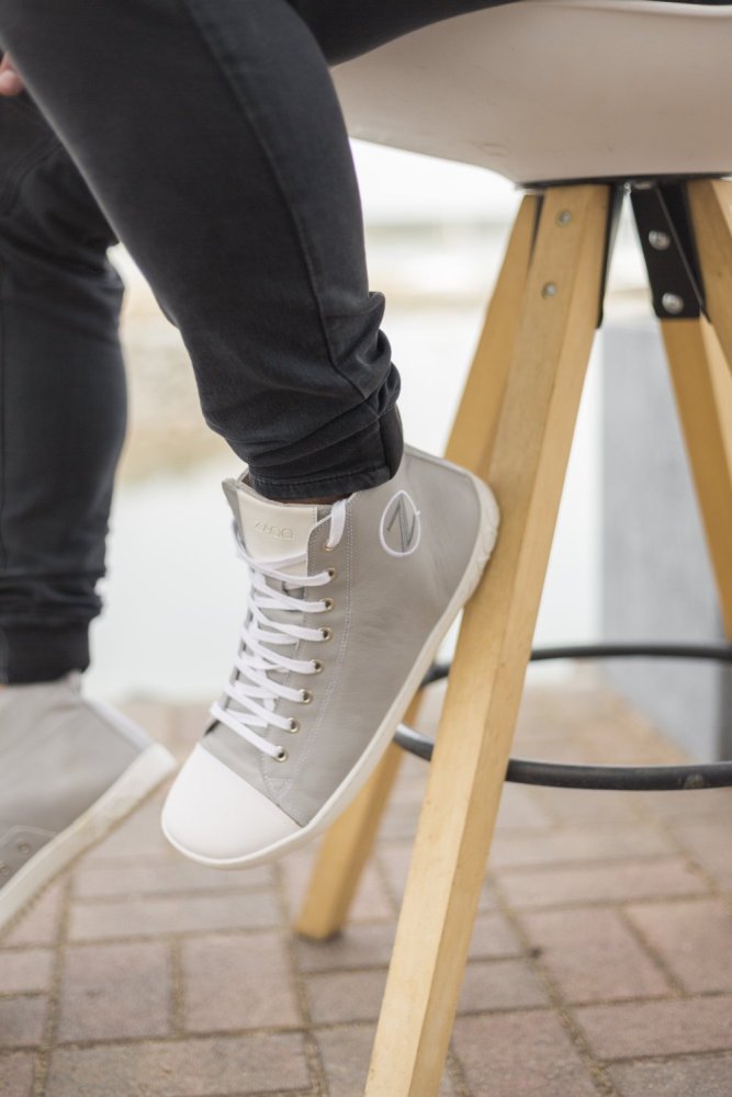 Eine Person, die ZAQQs CHUQQS Grey High-Top-Sneaker und eine schwarze Hose trägt, sitzt auf einem Holzhocker mit schwarzen Metallakzenten. Der Fokus liegt auf den stilvollen Schuhen, die sorgfältig aus Nappaleder gefertigt wurden, vor einem subtil verschwommenen Outdoor-Hintergrund.