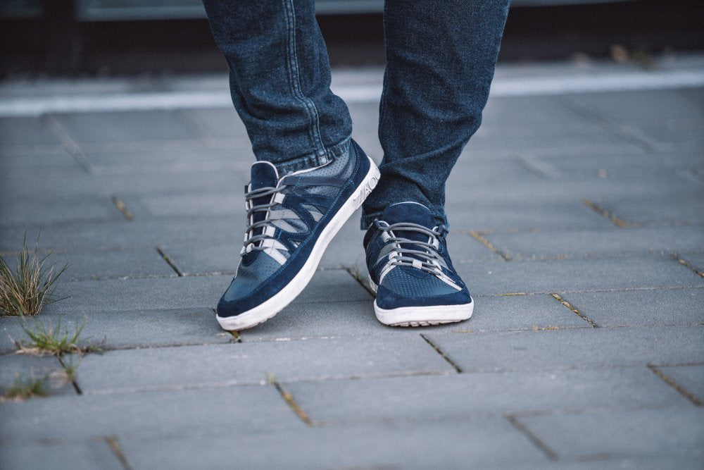 Eine Person in den für ihren Tragekomfort bekannten DRAQE Azul Sneakers von ZAQQ läuft auf einem grau gefliesten Gehweg. Die Person trägt Blue Jeans, zwischen den Fliesen lugt etwas Gras hervor.