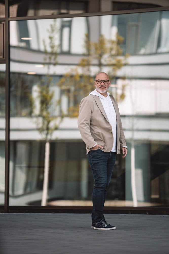 Ein Mann mit Bart, Brille, beigem Blazer, weißem Hemd, Jeans und DRAQE Azul-Schuhen von ZAQQ für optimalen Komfort steht im Freien vor einem modernen Glasgebäude, in dem sich Bäume spiegeln.