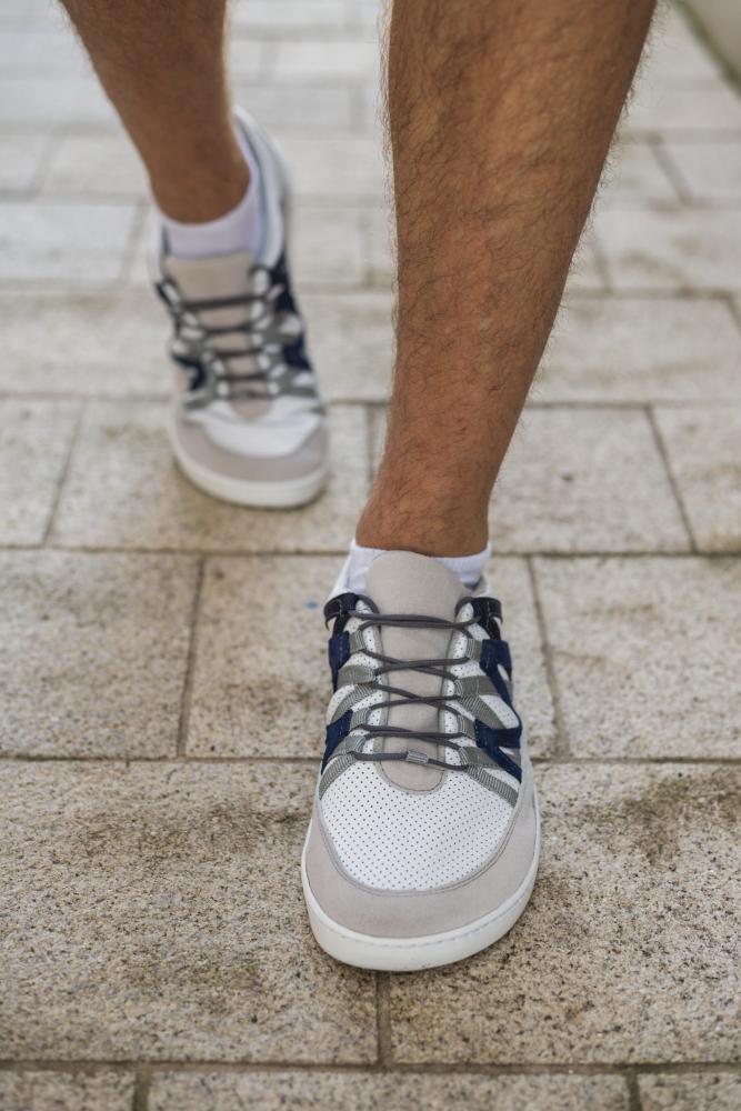 Eine Nahaufnahme einer Person, die auf einem gepflasterten Gehweg läuft und die DRAQE White Grey-Sneakers von ZAQQ mit schwarzen Schnürsenkeln trägt. Diese Schuhe aus hochwertigem Nappaleder werden mit weißen Socken kombiniert und fangen die lässige Eleganz des Barfußschuh-Stils perfekt ein.