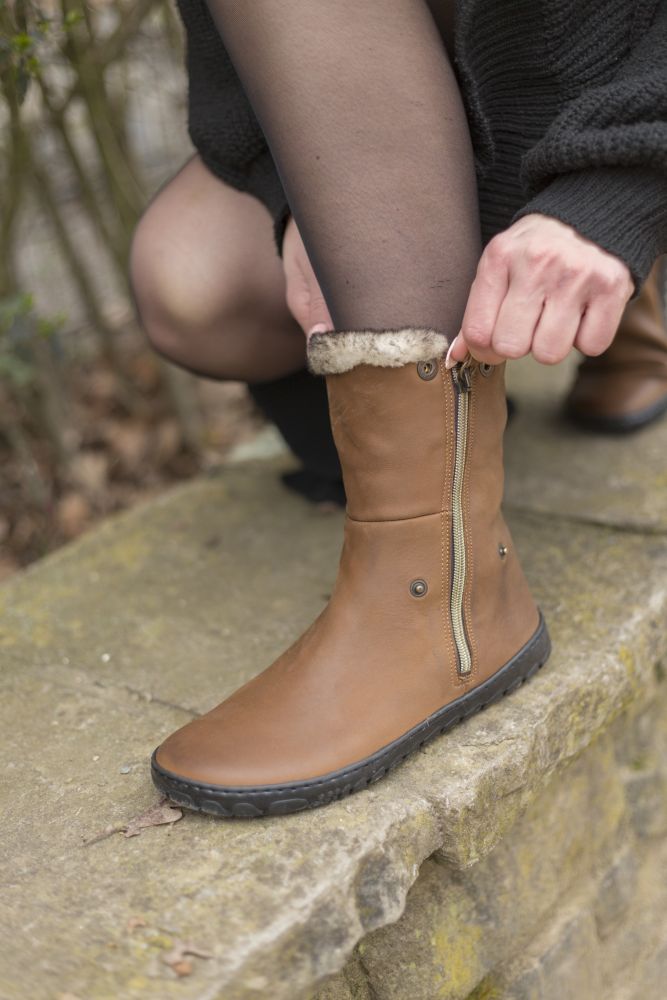 Eine Person in schwarzer Strumpfhose und schwarzem Mantel passt den HUQ Brown Boot von ZAQQ mit seitlichem Reißverschluss und Fellfutter aus Veloursleder an und sitzt dabei auf einem Felsvorsprung. Der Hintergrund besteht aus verschwommenem Laub und Steinen.