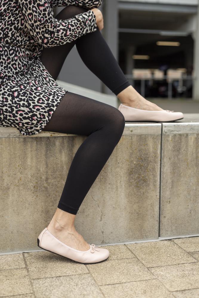 Eine Person sitzt gemütlich auf einem Betonvorsprung und trägt ein Kleid mit Leopardenmuster, schwarze Leggings und LOOQ Nude-Schuhe von ZAQQ. Das Outfit strahlt vor der Kulisse eines städtischen Außenbereichs mühelose Eleganz aus.