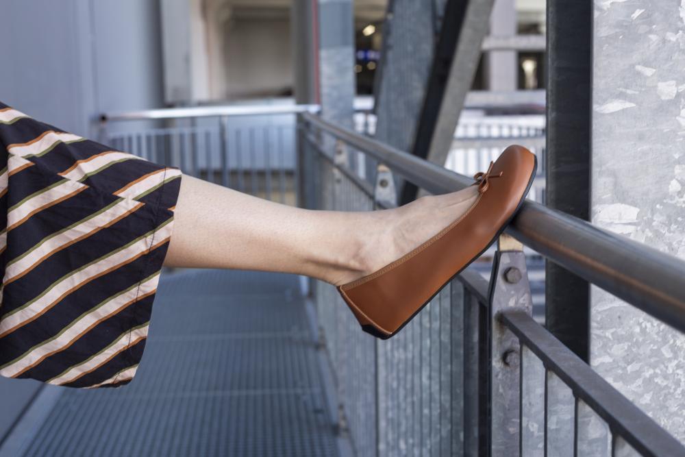 Eine Person trägt ein Paar braune flache Schuhe aus der LOOQ Vegan Cognac-Kollektion von ZAQQ und streckt ihr Bein auf einem Metallgeländer aus. Sie trägt Hosen mit schwarzen und beigen Streifen vor dem Hintergrund industrieller Metallstrukturen.