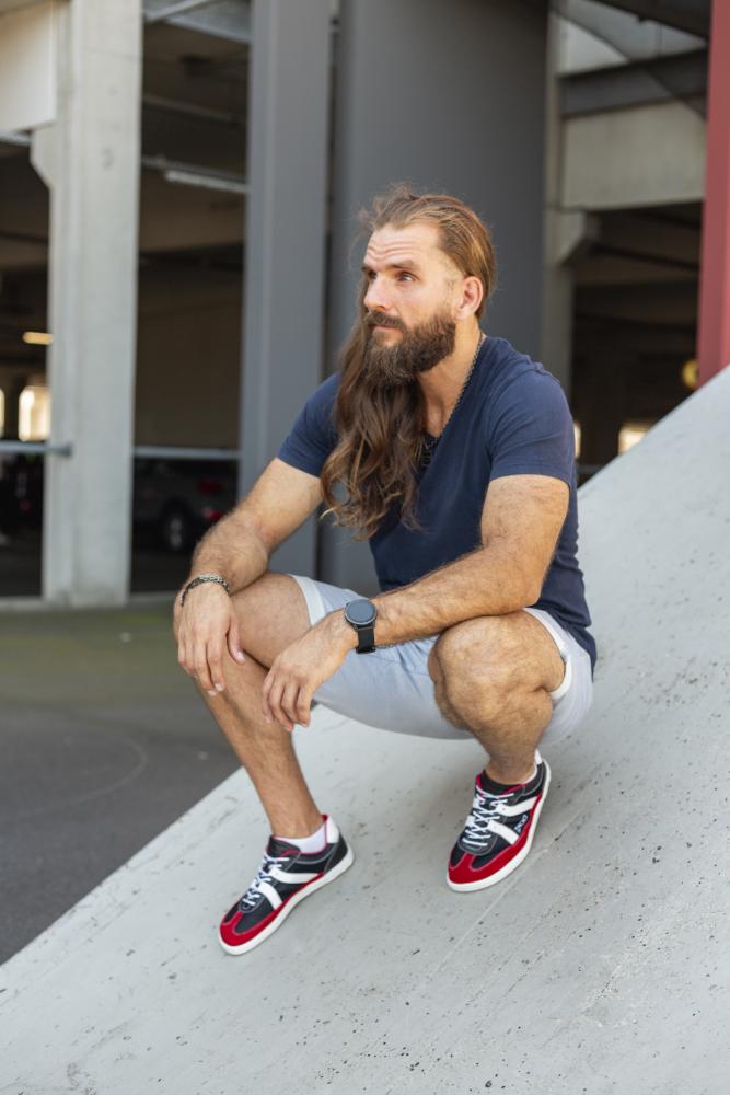 Ein bärtiger Mann mit langen Haaren hockt auf einer Betonrampe. Er trägt ein marineblaues T-Shirt von OAQLEY, hellgraue Shorts und auffällige rote ZAQQ-Sneaker. Im Hintergrund ist das schwach beleuchtete Innere eines Parkhauses zu sehen.