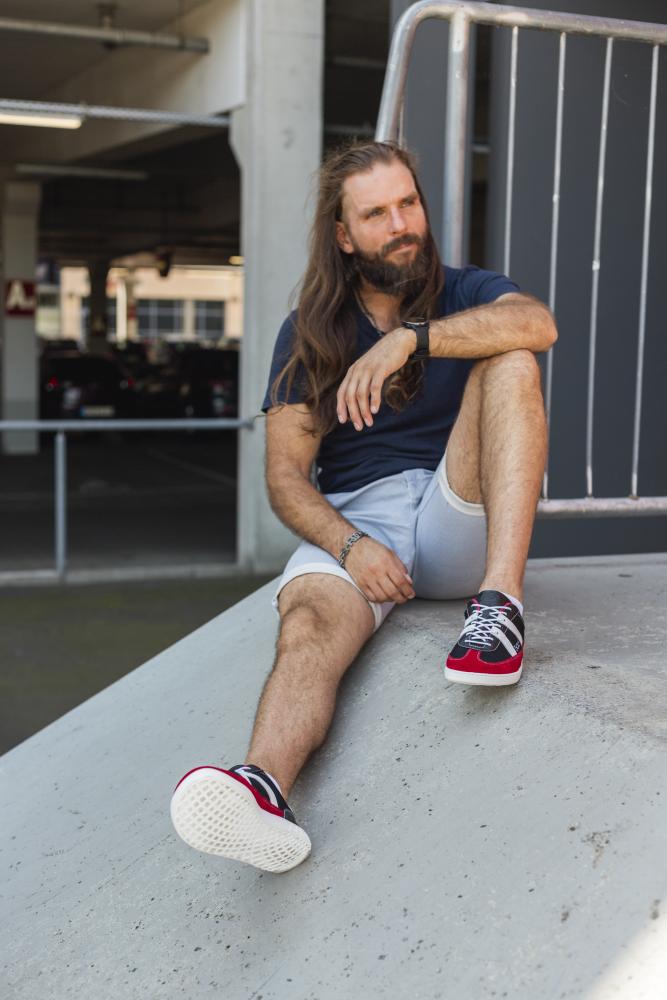 Ein Mann mit langen Haaren und Bart sitzt lässig auf einer Betonfläche auf einem Außenparkplatz. Er trägt ein dunkles Hemd, helle Shorts und rote OAQLEY-Sneaker von ZAQQ.