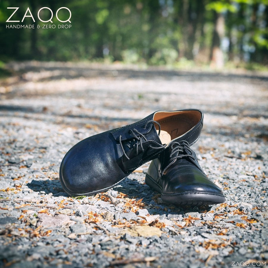 Die PEAQ Black-Schuhe von ZAQQ stehen auf einem Schotterweg inmitten eines Waldes. Diese schwarzen Lederschuhe haben ein elegantes „Leder“-Finish und zeichnen sich durch ein minimalistisches, bequemes Design aus. Sonnenlicht fällt durch die Bäume und beleuchtet die Szene mit dem Markentext: ZAQQ Handmade & Zero Drop.