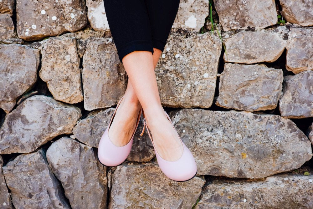 Eine Person in ZAQQs PINQ Candy Ballerinas, einem eleganteren Sommerschuh und einer schwarzen Hose sitzt mit gekreuzten Beinen auf einer Steinmauer. Die raue Textur der Steine kontrastiert mit der Glätte dieser femininen, zarten Schuhe.