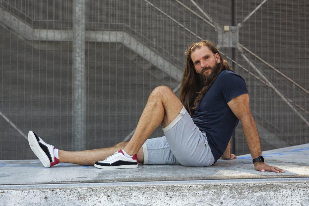 Ein Mann mit langen Haaren, einem blauen T-Shirt, hellgrauen Shorts und Barfuß-Sneakern QAMPION Black White von ZAQQ mit flexibler Sohle, sitzt mit einem ausgestreckten Bein auf einer Betonfläche. Er befindet sich im Freien, im Hintergrund sind Metalltreppen und ein Zaun zu sehen.