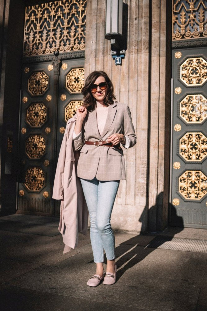 Eine Frau mit Sonnenbrille posiert selbstbewusst vor einer verzierten Tür mit goldenen Details und zeigt ihren eleganten Stil. Sie trägt einen hellen Blazer und einen passenden Mantel, dazu hellblaue Jeans und rosa Schuhe mit einer glatten Kautschuksohle aus der QERP Candy-Linie der Marke ZAQQ.