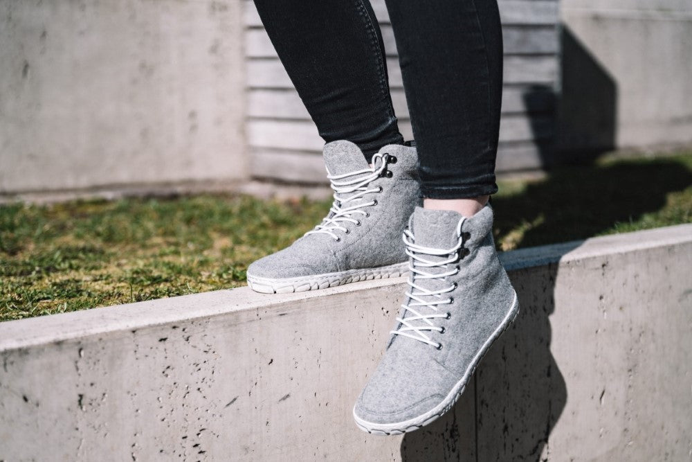 Eine Person, die die ZAQQ QICE Grey High-Top-Sneaker mit weißen Schnürsenkeln und einer Outdoor-Sohle trägt, sitzt auf einem Betonvorsprung. Sie ist mit schwarzen Hosen bekleidet, im Hintergrund sind Gras und ein Gebäude zu sehen.