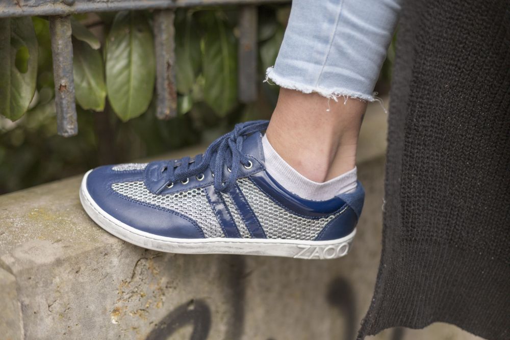 Nahaufnahme einer Person, die die blau-weißen QIMBERLY Blue-Sneakers von ZAQQ mit Mesh-Details trägt und auf einer Betonkante steht. Die Person trägt Bluejeans mit ausgefransten Säumen und ein schwarzes Kleidungsstück. Im Hintergrund sind grüne Blätter zu sehen.