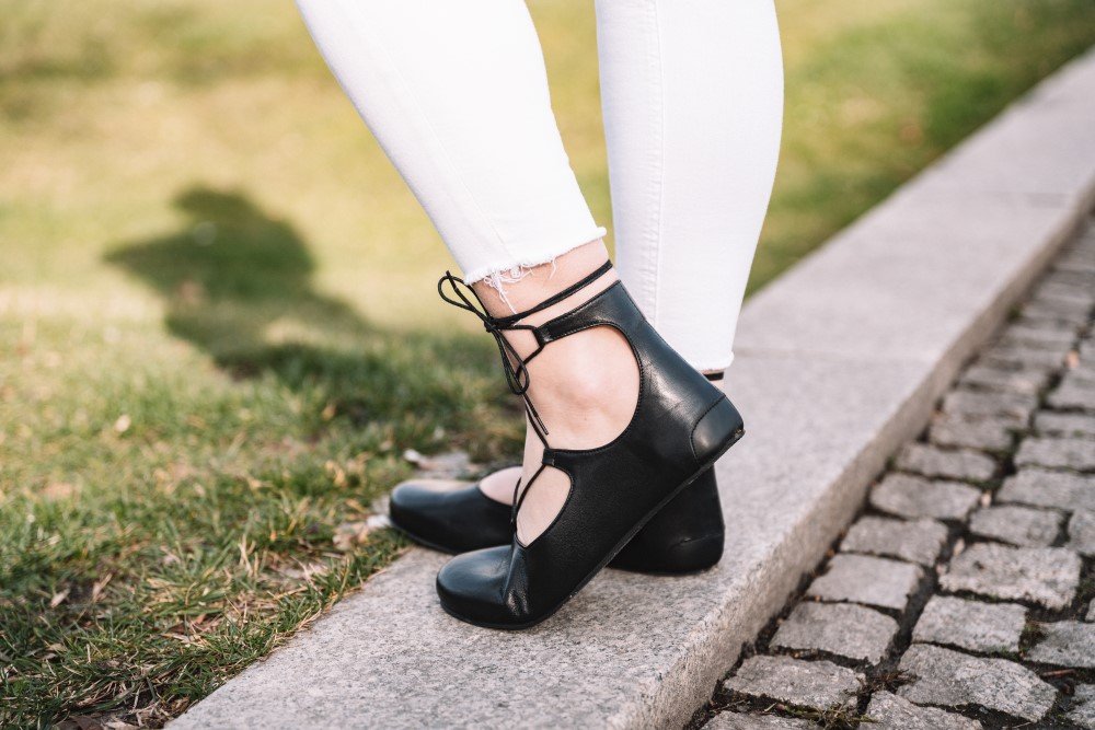 Eine Person, die schwarze Ballerinas und weiße Hosen von ZAQQ trägt, steht auf einem Steinbordstein. Das Foto zeigt ihre Füße und Unterschenkel vor einem Hintergrund aus Gras und Kopfsteinpflaster, wobei die Schatten elegant auf den Boden geworfen werden.