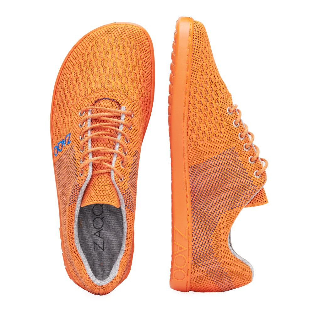 Wir präsentieren die QNIT Orange Sneaker von ZAQQ: Diese leuchtend orangefarbenen Schuhe sind mit blauen Akzenten versehen, weisen eine atmungsaktive Mesh-Textur auf und verfügen über ein bequemes Barfußschuh-Design. Ein Sneaker ist von oben zu sehen, während der andere von der Seite gezeigt wird. Sowohl an der Seite als auch auf der Innenseite ist der Markenname ZAQQ deutlich zu erkennen.