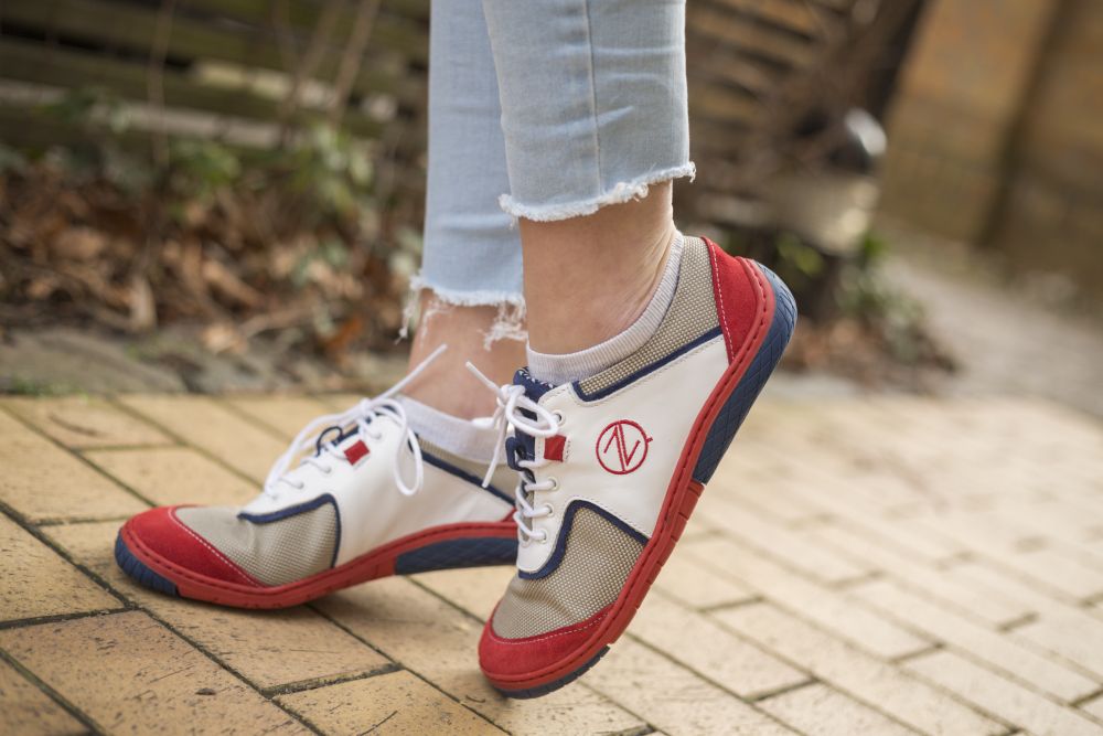 Eine Person in hellblauen Jeans mit ausgefranstem Saum und den weißen Sneakers QOLETTE der Marke ZAQQ steht auf einem gepflasterten Weg. Die Sneakers tragen an der Seite ein Logo mit dem Buchstaben V in Rot, Weiß und Blau.