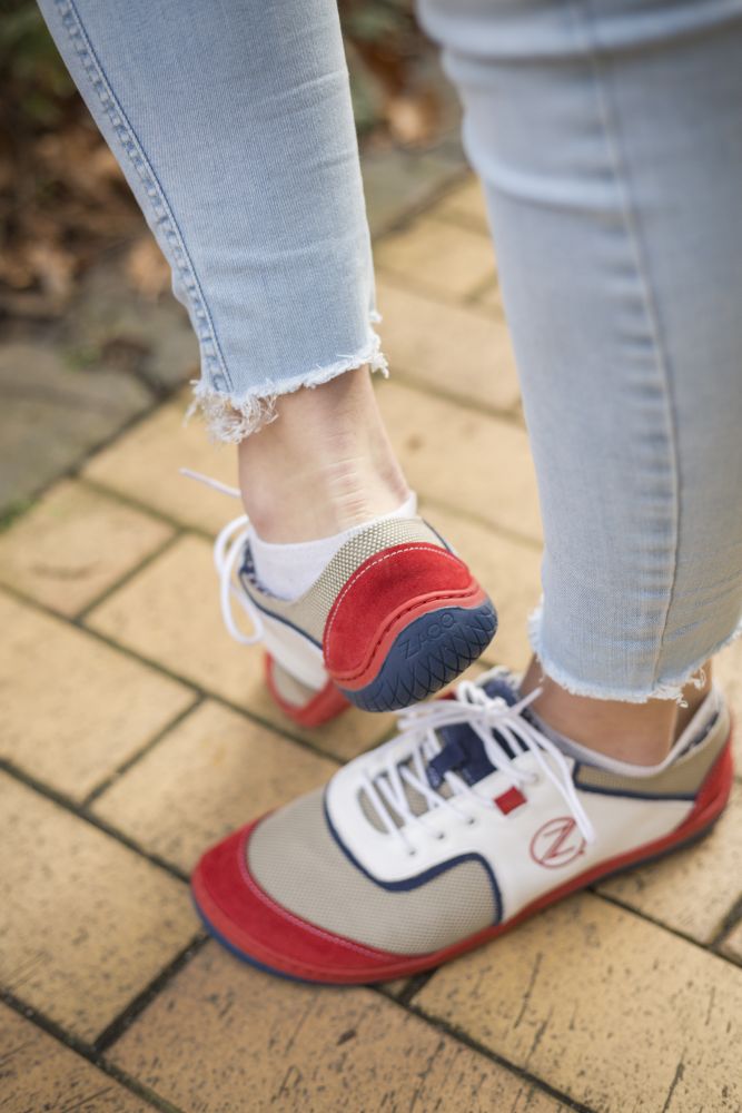 Eine Nahaufnahme zeigt eine Person in hellblauen zerrissenen Jeans und den weißen ZAQQ QOLETTE-Sneakern mit roten und blauen Details, die auf einem gelben Ziegelweg steht. Ein Fuß ist leicht angehoben, sodass die Sohle des Schuhs sichtbar ist, während trockene Blätter den Weg säumen und eine Stimmung der ZAQQ Kollektion einfangen.