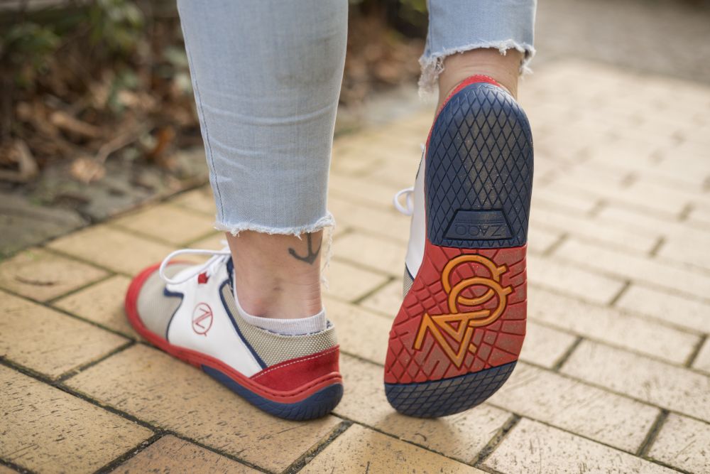 Eine Person in hochgekrempelten hellblauen Jeans läuft einen Ziegelweg entlang und präsentiert ihre weißen QOLETTE-Sneaker von ZAQQ. Über einem Knöchel lugt ein Tattoo hervor, während die Sohle des Schuhs ein markantes Design mit einem Logo aufweist.