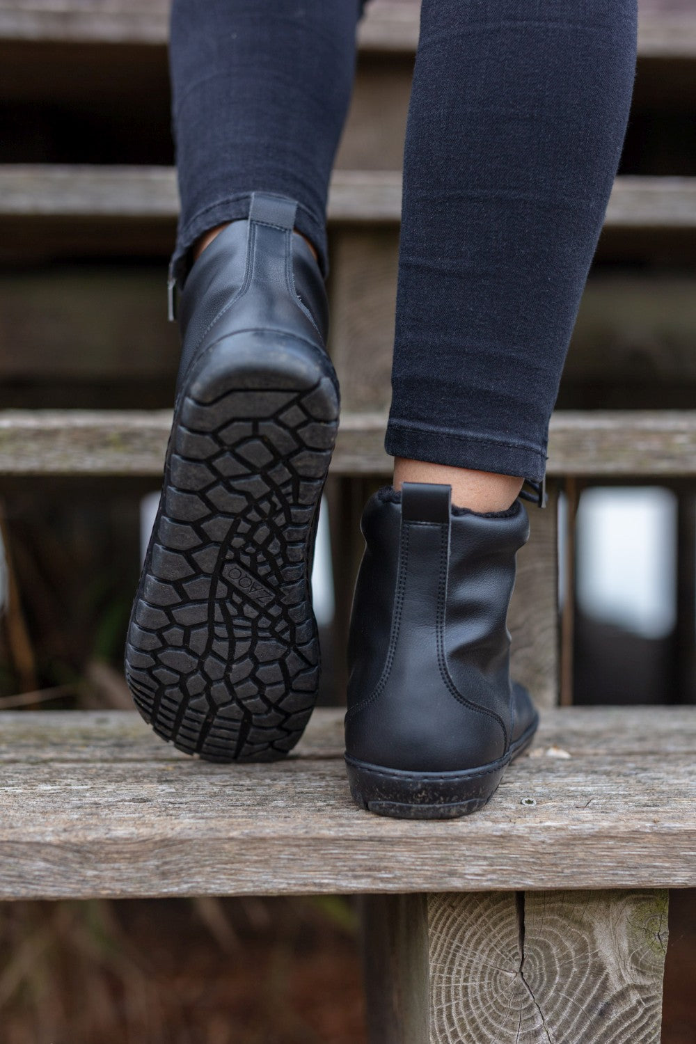 Das Nahaufnahmebild zeigt eine Person, die Holzstufen hinaufsteigt. Die dunkle Röhrenjeans und die QUINTIC Winter Vegan Black Stiefeletten von ZAQQ sind besonders gut zu erkennen. Diese Stiefel sind mit strukturierten Gummisohlen mit einzigartigem Muster ausgestattet und bieten außergewöhnliche Haltbarkeit und Halt. Der leicht unscharfe Hintergrund betont die wasserabweisende Verarbeitung des Schuhs.