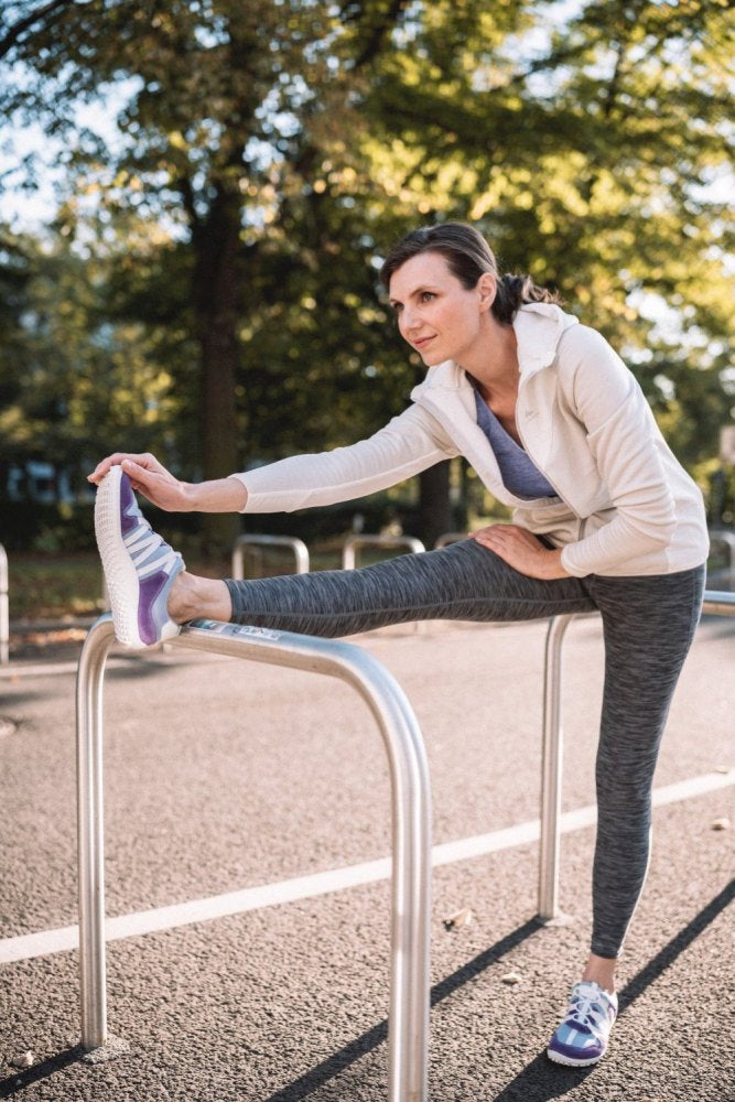 Eine Frau in Sportkleidung streckt ihr Bein auf einem Metallgeländer in einem Park aus. Sie trägt eine weiße Jacke, graue Leggings und RIQ Purple Sneakers von ZAQQ aus atmungsaktivem Meshgewebe. Im Hintergrund sind Bäume zu sehen, die in der Sonne liegen.