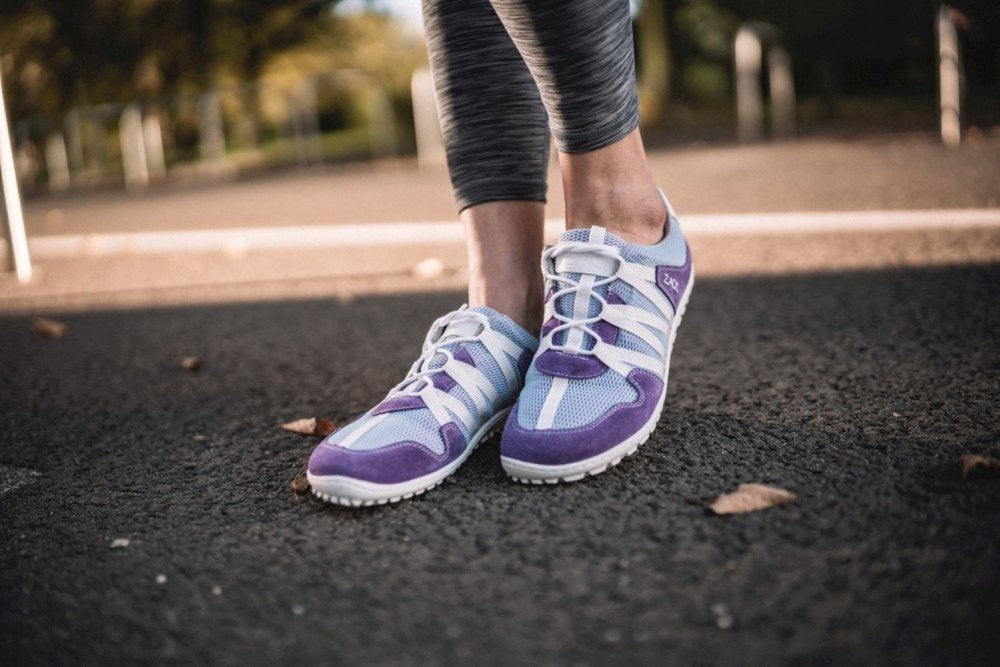 Nahaufnahme einer Person, die ZAQQs RIQ Purple-Sneaker aus Meshgewebe trägt und auf einem Asphaltweg mit verstreuten Blättern steht. Der Hintergrund zeigt eine verschwommene Ansicht von Bäumen und Masten, was auf einen Park oder eine Außenumgebung hindeutet.