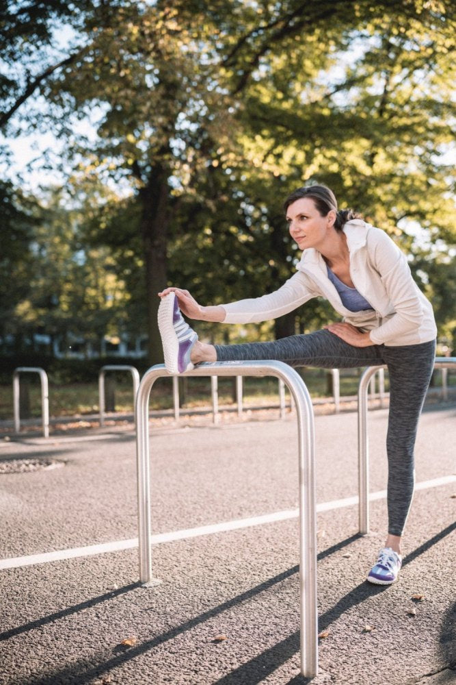 Eine Frau in Sportkleidung streckt ihr Bein auf einem Metallgeländer in einem Park aus. Sie trägt eine leichte Jacke, Leggings und RIQ Purple Barfußschuhe von ZAQQ. Im Hintergrund sind Bäume und ein sonniger Tag zu sehen.