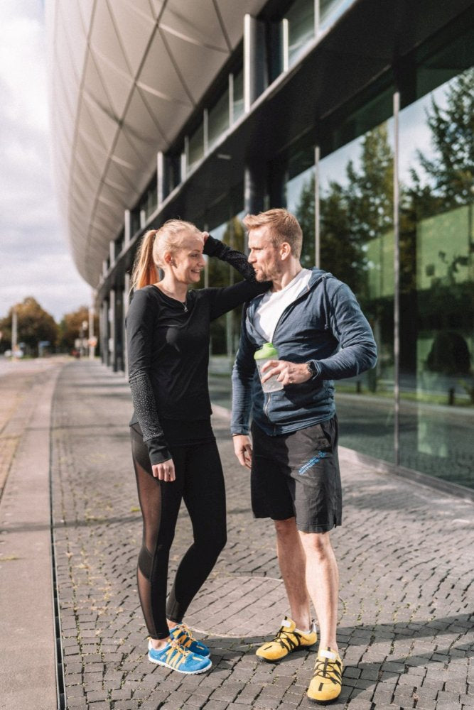 Ein Mann und eine Frau in Sportkleidung stehen vor einem modernen Gebäude mit Glasfenstern. Die Frau, deren Haare zurückgebunden sind, lächelt, während der Mann ein Getränk in der Hand hält. Beide wirken entspannt in ihren ZAQQ RIQ Blue Orange-Schuhen, während sie sich auf dem Kopfsteinpflaster unterhalten.