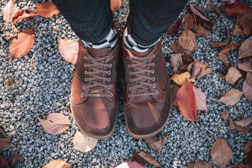 Der ZAQQ RIQO Brown sorgt in Kombination mit gestreiften Socken für außergewöhnlichen Komfort auf einem Kiesweg, der mit verstreuten Herbstblättern geschmückt ist.