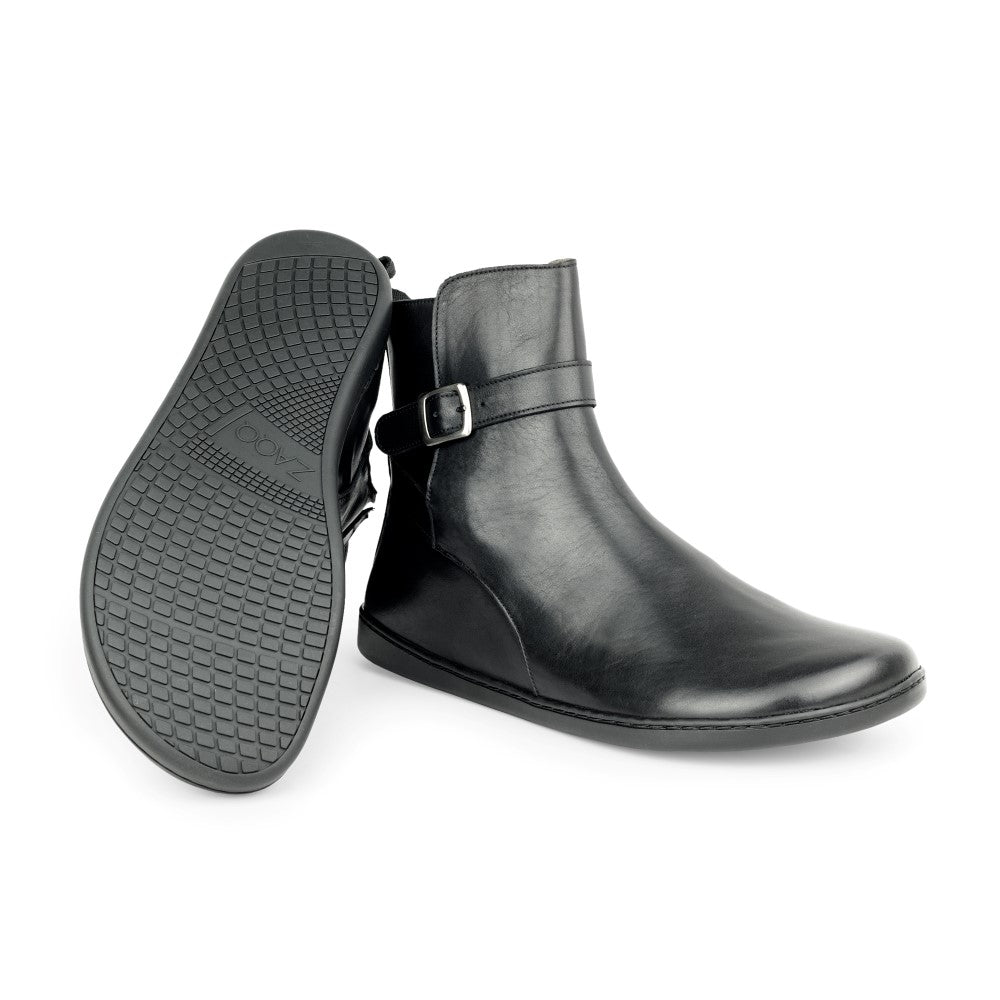 Ein Paar schwarze RIQUET-Stiefeletten von ZAQQ, eine steht aufrecht, die andere zeigt das Profilmuster der Sohle. Diese eleganten Stiefel haben eine seitliche Schnalle und ein elegantes Aussehen, das sowohl Stil als auch Komfort bietet.