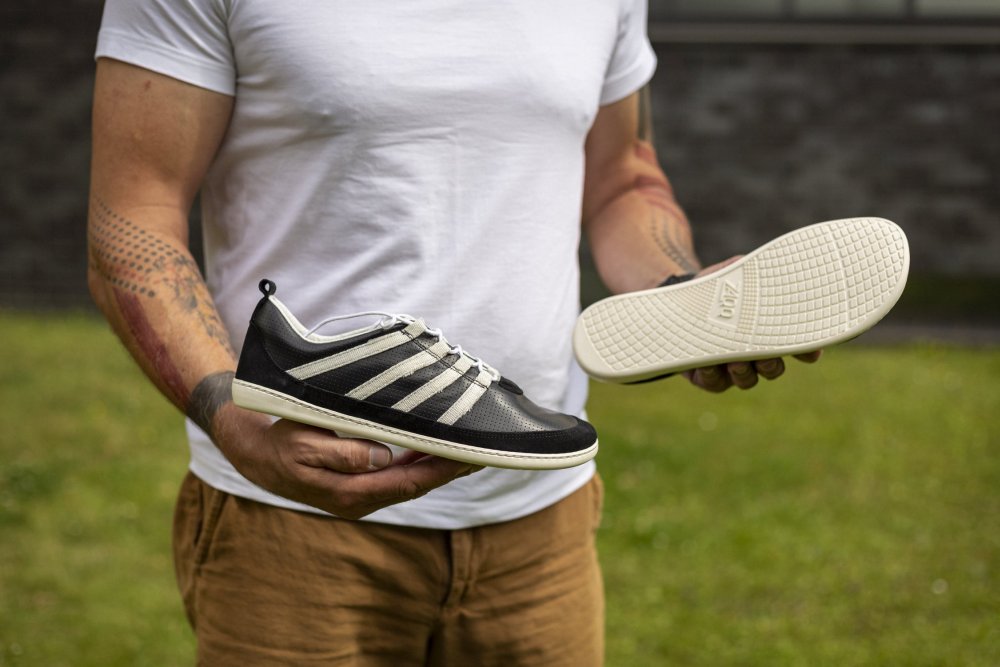 Auf einer Rasenfläche mit einer dunklen Backsteinmauer im Hintergrund hält eine Person, bekleidet mit einem weißen Hemd und braunen Hosen, ein Paar SPARQ Low Black White-Sneaker von ZAQQ, wobei sowohl die Sohle als auch die Seitenansicht im Freien deutlich zu sehen sind.