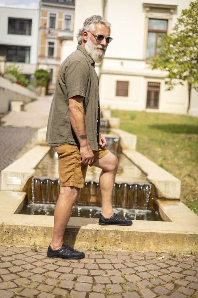 Ein Mann mit Bart und Sonnenbrille steht selbstbewusst mit einem Fuß auf dem Rand eines Steinbrunnens. Er trägt ein grünes Hemd, braune Shorts und stylische SPARQ Low Black Sneakers von ZAQQ. Im Hintergrund sind ein Gebäude und Grünflächen zu sehen.