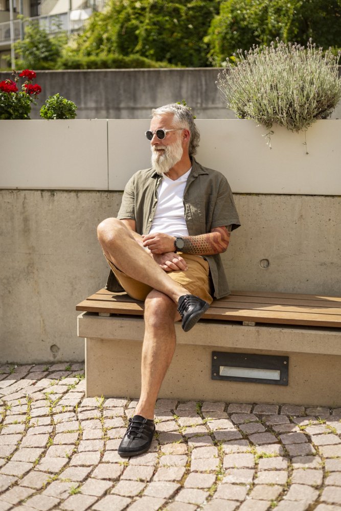 Ein bärtiger Mann mit Sonnenbrille, weißem T-Shirt, grünem Hemd und braunen Shorts sitzt auf einer Holzbank in einer sonnigen Umgebung im Freien. Er sitzt entspannt mit gekreuzten Beinen da und präsentiert seine stylischen SPARQ Low Black Sneakers von ZAQQ. Üppige Pflanzen und leuchtende Blumen schaffen eine ruhige Kulisse.