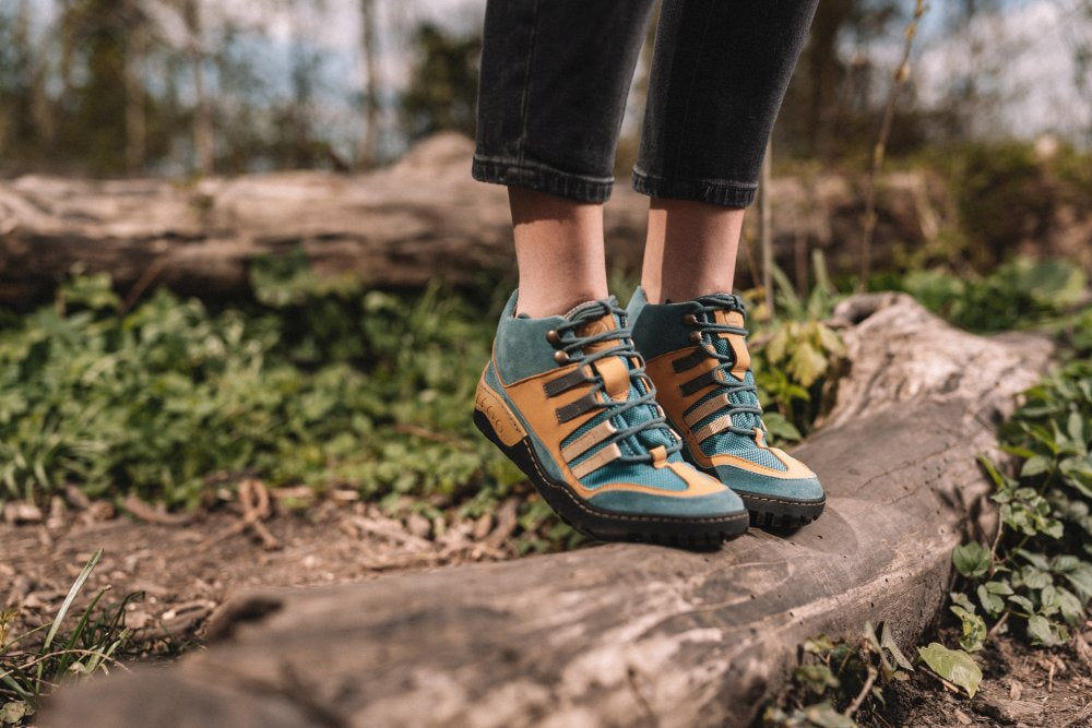 Eine Person, die die leuchtenden SQOUT Blue Orange Waterproof-Stiefel von ZAQQ trägt, steht auf einem umgestürzten Baumstamm inmitten üppiger grüner Blätter. Die Szene weckt ein Gefühl von Abenteuer und Erkundung im Freien und lädt Sie ein, die Erheiterung mit atmungsaktivem Komfort zu genießen.