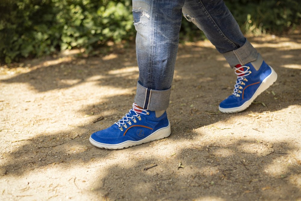 Eine Person in hochgekrempelten Jeans und einem Paar TERRAQ Azure Waterproof-Sneakers mit weißen Schnürsenkeln von ZAQQ schlendert einen Feldweg entlang. Die Szene spielt im Freien, inmitten üppigen Grüns und gesprenkeltem Sonnenlicht.