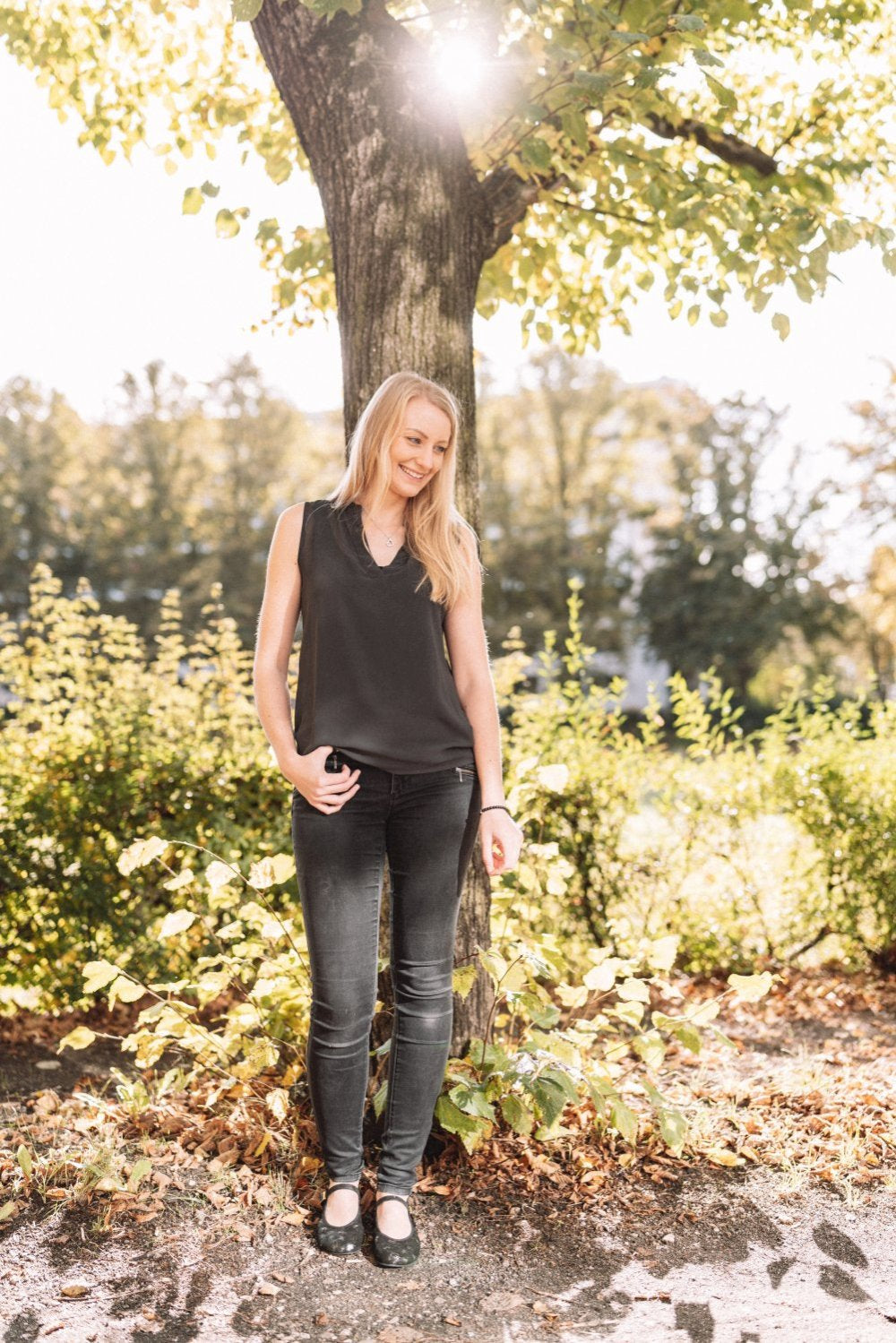 Eine Frau mit langen blonden Haaren steht lächelnd unter einem Baum in einem sonnenbeschienenen Park und strahlt in ihrem schwarzen ärmellosen Top und dunklen Jeans Eleganz aus, stilvoll ergänzt durch ZAQQs TWIST Ennis Ballerinas. Der Hintergrund ist mit grünem Laub gefüllt, das vom Sonnenlicht beleuchtet wird.